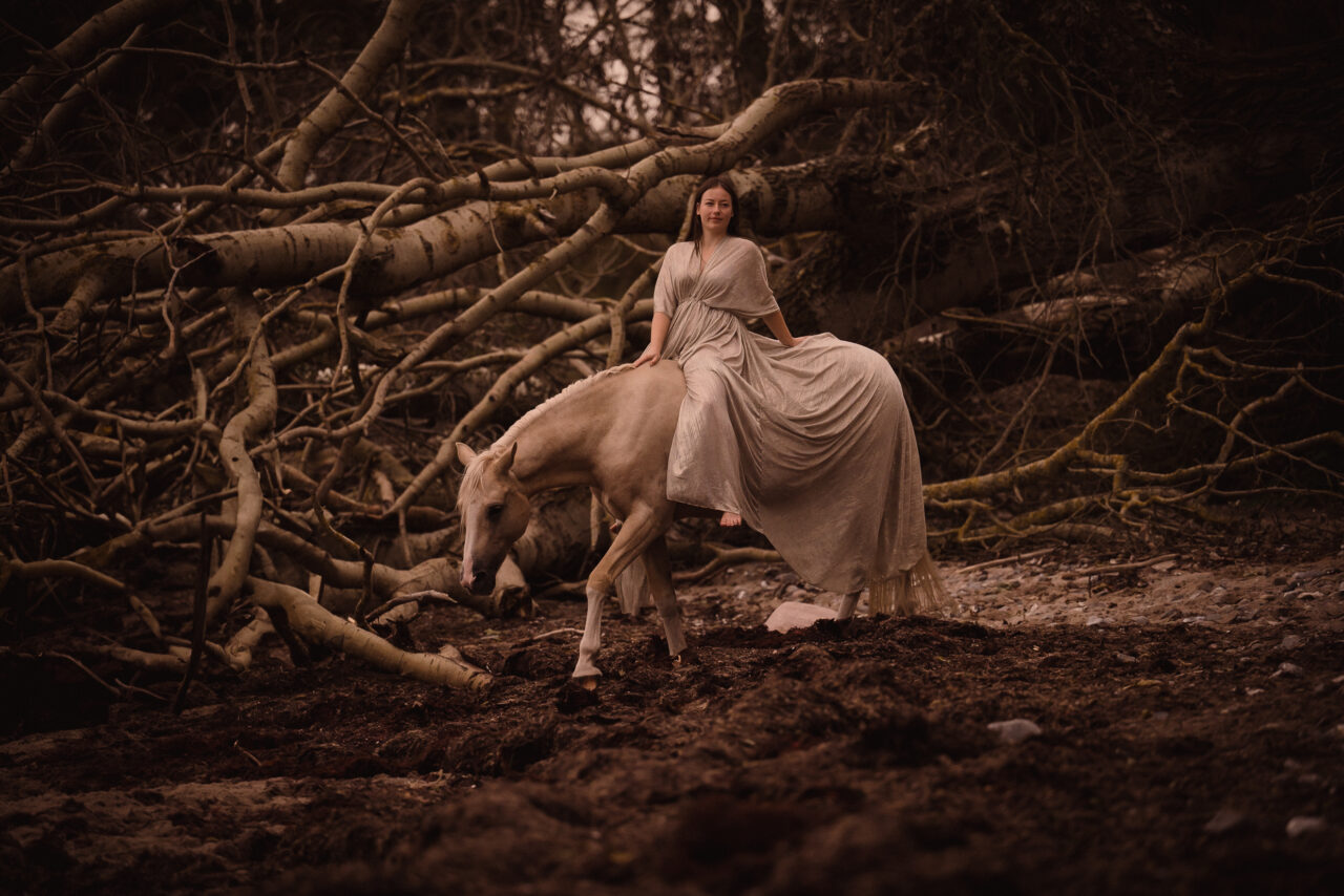 Portrait einer Frau in einem langen weißen Kleid, auf einem weißen Pferd sitzend, in einer Strandlandschaft mit umgestürzten Bäumen