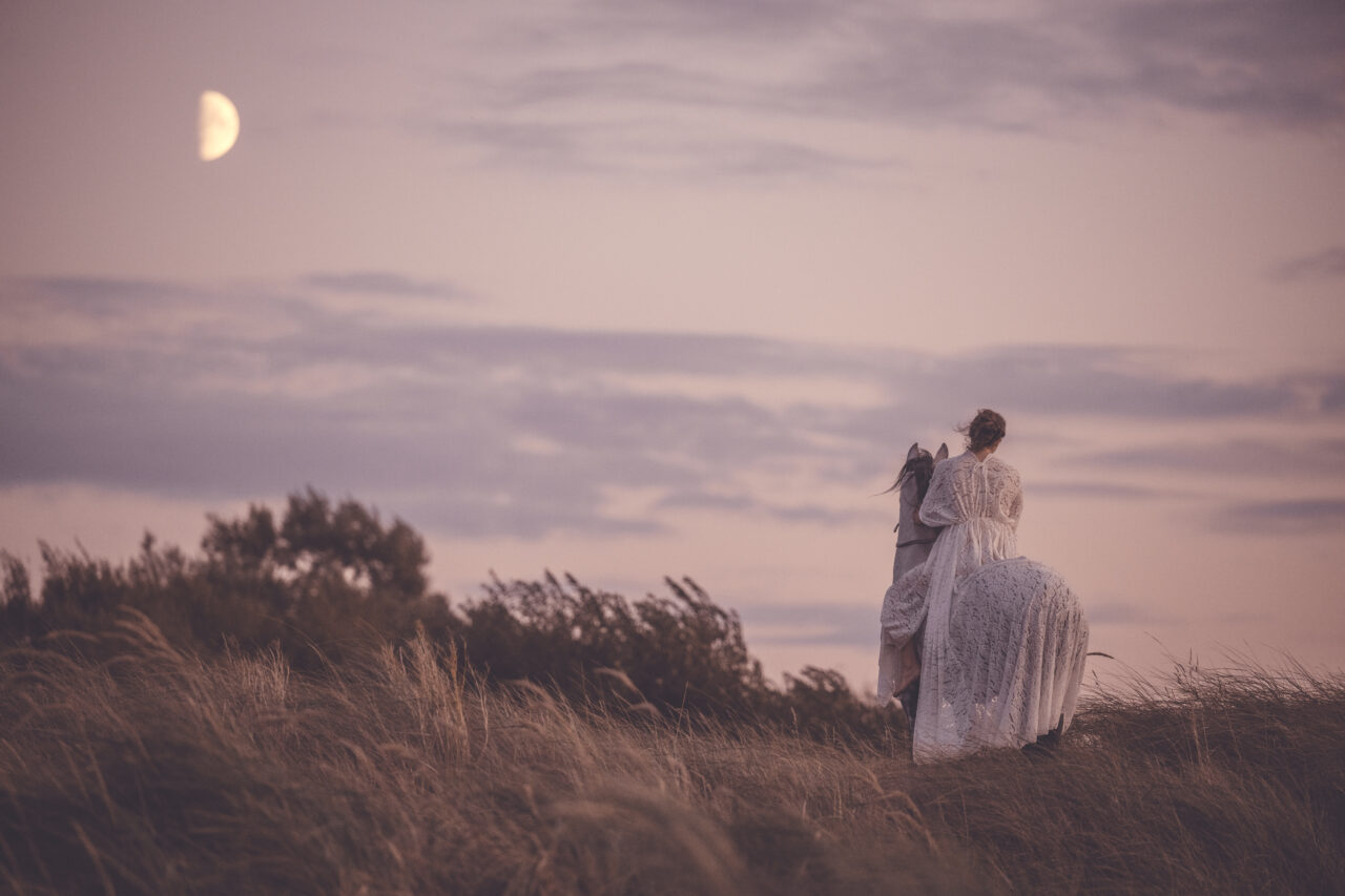 Frau mit weißem langen Kleid auf einem Pferd vor einem Sonnenuntergang mit Halbmond